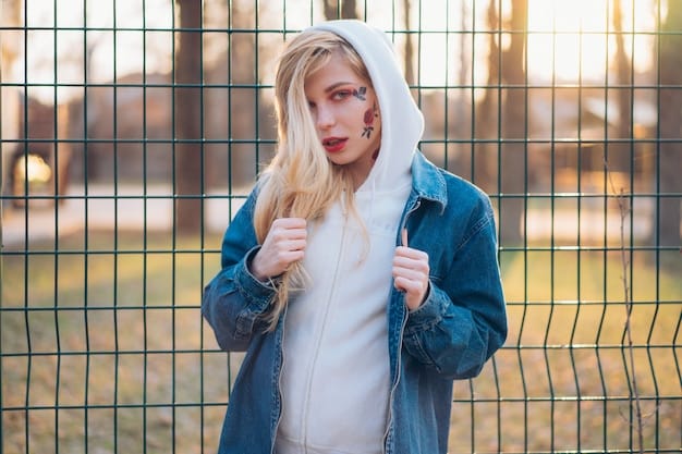 A young woman wearing a large, colorful oversized hoodie, baggy ripped jeans, and chunky white sneakers, posing in front of a graffiti-covered wall. She wears a backwards baseball cap and has brightly colored hair.
