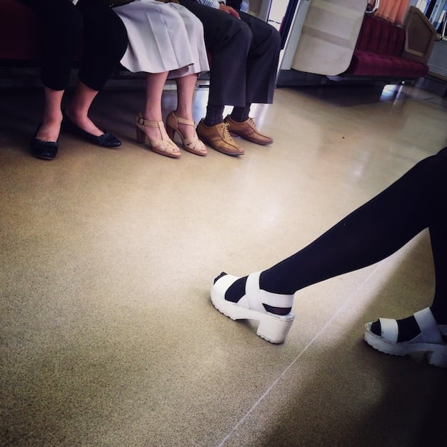 A close-up shot of a person's feet as they follow dance steps in a K-Pop tutorial. The focus is on the precise footwork and the instructor's feet in the background.