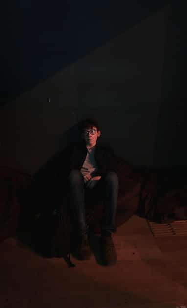 A still from a K-drama showing the main character sitting alone in a dimly lit room, looking despondent. The character is surrounded by stacks of books, suggesting a sense of isolation and being overwhelmed. The lighting is soft and melancholic, enhancing the feeling of sadness and introspection.